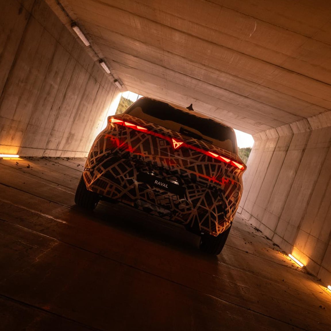 De CUPRA Raval, bedekt met een bruin-witte stadsplattegrond camouflage wrap, staat binnen tentoongesteld. Achter de auto bevindt zich een hekwerkpaneel met een oplichtend wit CUPRA logo en de tekst "BEYOND REBEL, RAVAL.".