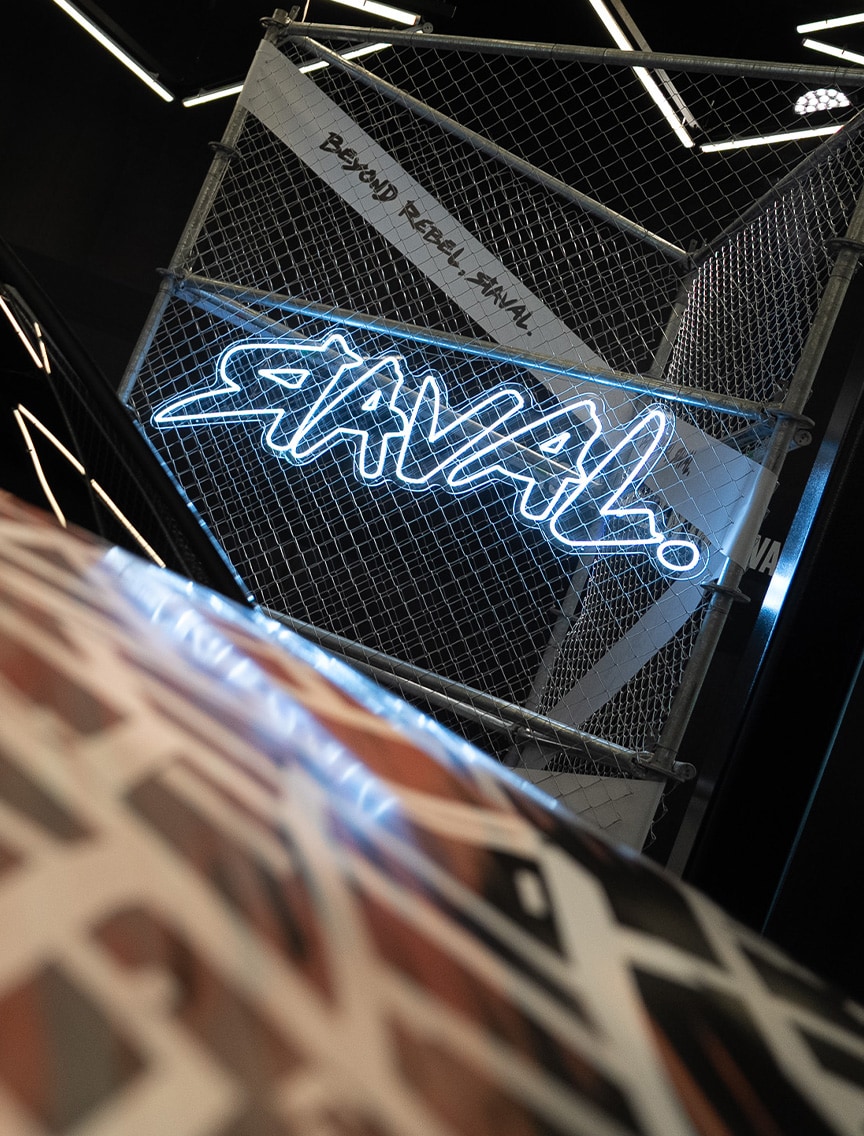 Een neonlicht dat "RAVAL!" spelt, is weergegeven op een hekwerkpaneel. Dit paneel bevat ook de tekst "BEYOND REBEL, RAVAL.". Het geheel staat in een donkere, gestileerde ruimte met geometrische lichtpatronen.