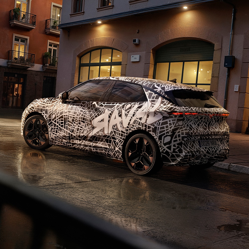 Een zij- en achteraanzicht van de CUPRA Raval. De auto is bedekt met een zwart-witte camouflagewrap met daarop een stadsplattegrond, en met de grote, gestileerde tekst "RAVAL" zichtbaar op de zijkant.