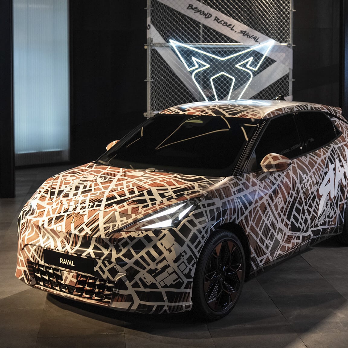De CUPRA Raval, bedekt met een bruin-witte stadsplattegrond camouflage wrap, staat binnen tentoongesteld. Achter de auto bevindt zich een hekwerkpaneel met een oplichtend wit CUPRA logo en de tekst "BEYOND REBEL, RAVAL.".